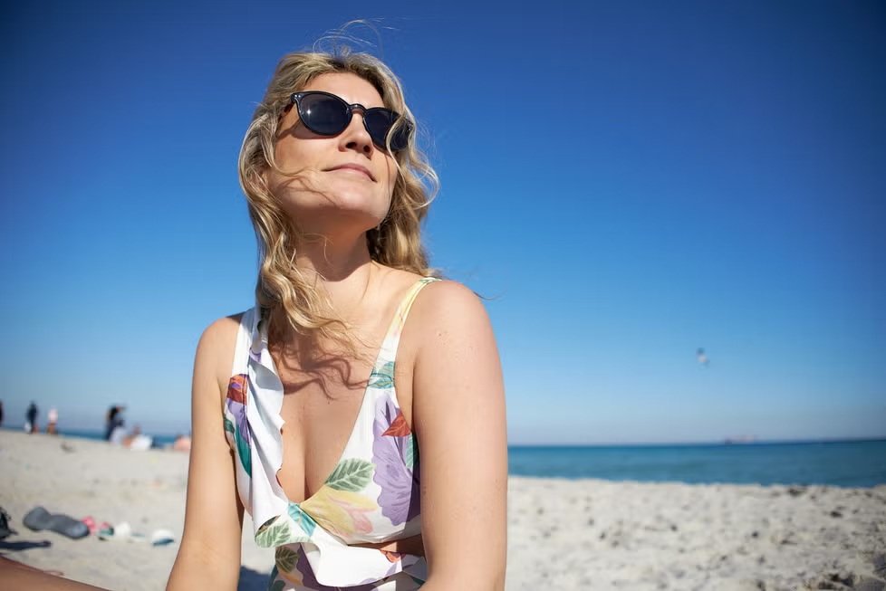 Mujer en la playa con gafas de sol, expuesta al sol y protegiendo su piel frente al daño solar y el envejecimiento cutáneo.